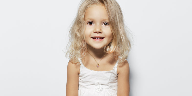 Lächelndes blondes Mädchen in weißem Kleid trägt zarte Halskette mit kleinem Schutzengel-Anhänger