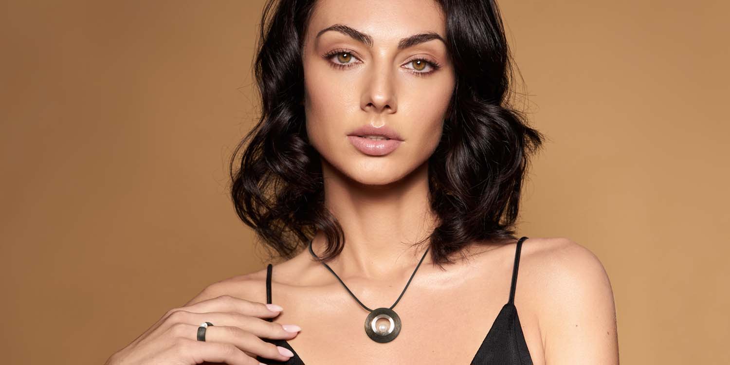 Elegante Frau mit dunklen Locken trägt schwarze Halskette mit rundem Anhänger und schwarzen Ring vor beigem Hintergrund