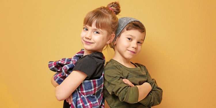 Zwei lächelnde Geschwister stehen Rücken an Rücken vor gelbem Hintergrund - Mädchen mit Dutt in kariertem Hemd, Junge mit Mütze in olivgrünem Pullover